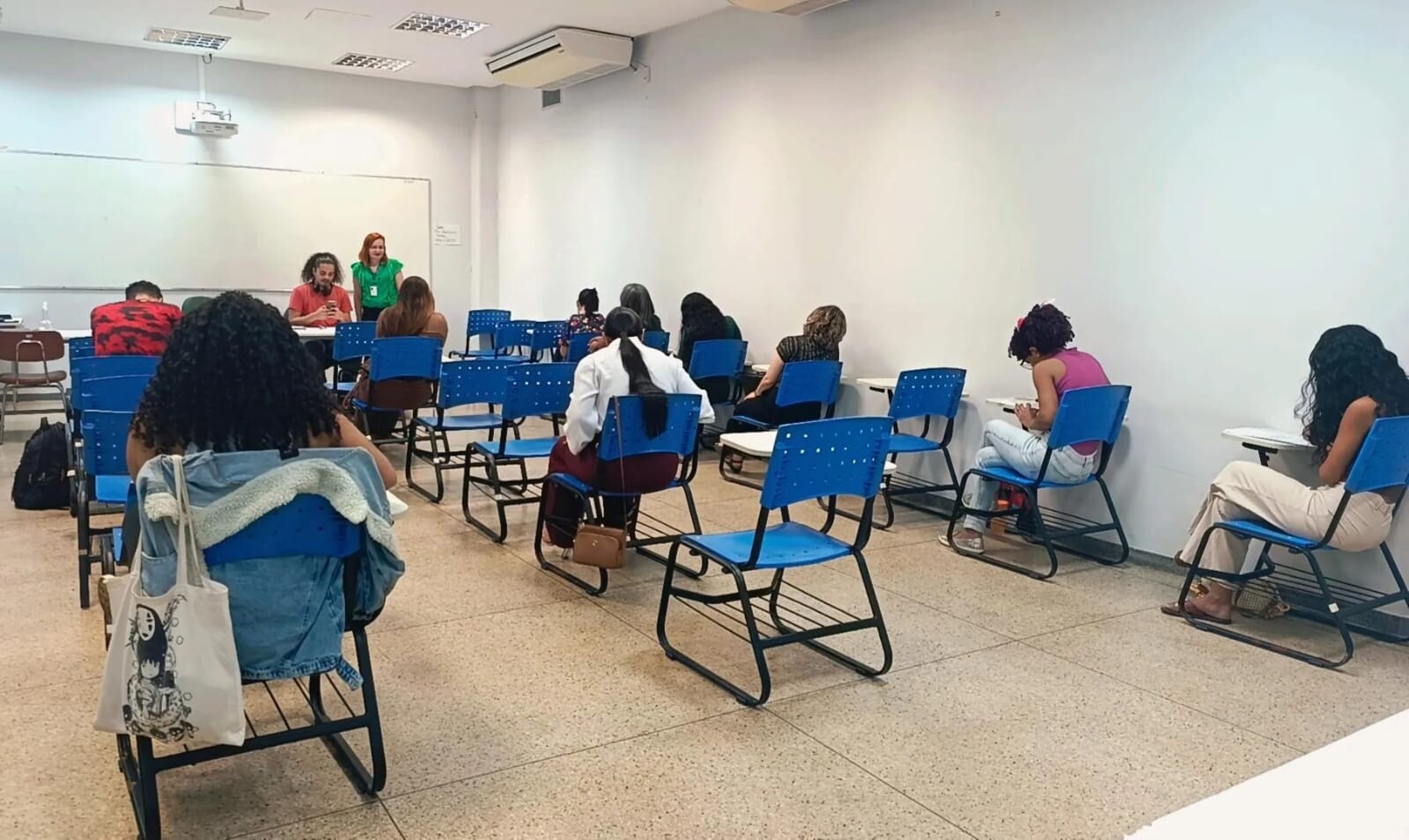 Foto de momento de realização de prova, com pessoas sentadas, de costas, escrevendo e duas pessoas em pé à frente em posição de fiscalização. 