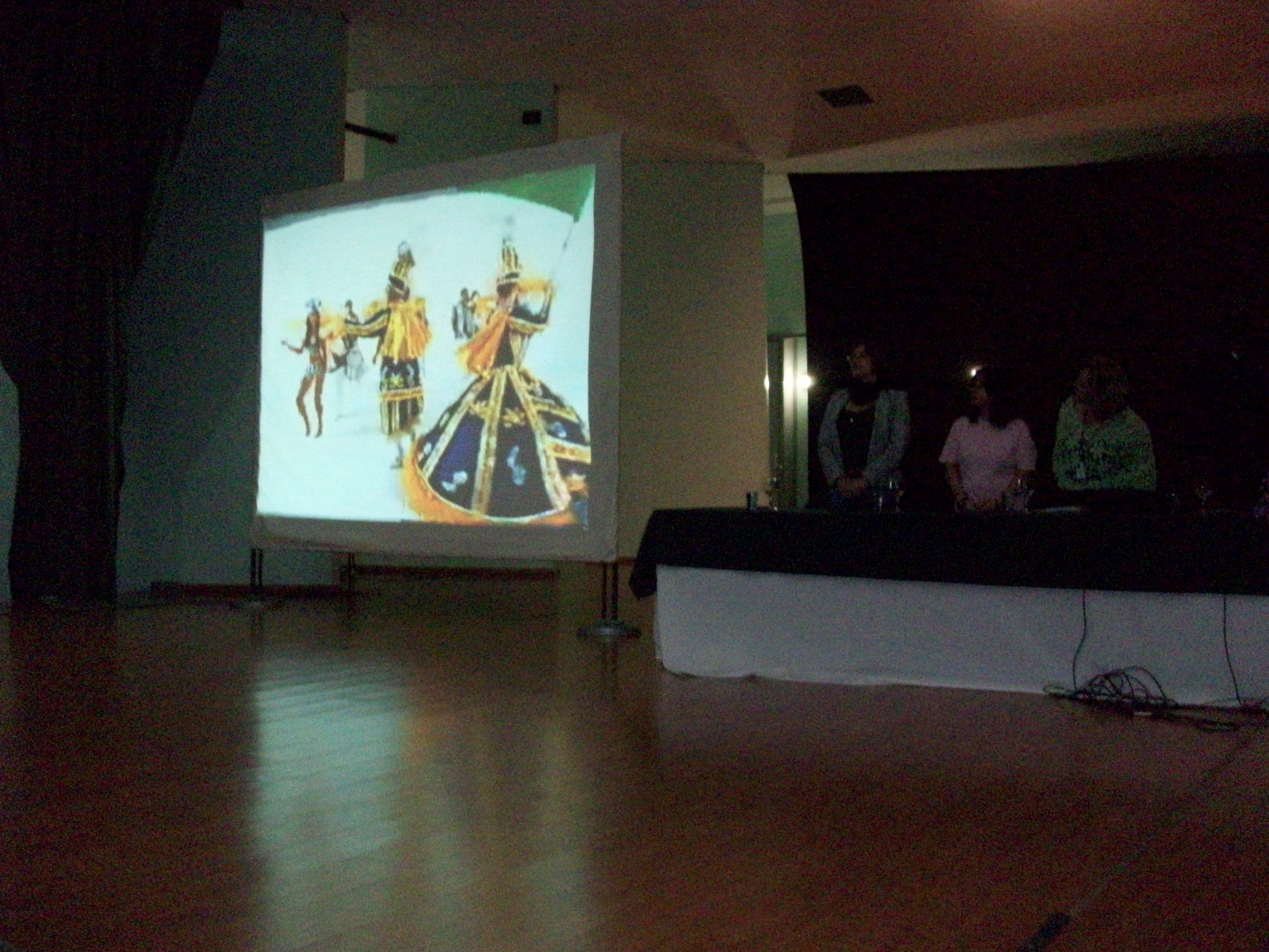 Professores na mesa do evento observando um slide 