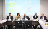 Cinco pessoas estão sentadas à mesa em uma reunião institucional, posicionadas à frente de um telão que exibe a frase "A INDÚSTRIA CRIA" e um QR Code. Os participantes, homens e uma mulher, vestem trajes formais e casuais, interagindo em um ambiente profissional com microfones e copos sobre a mesa. No centro da projeção, destaca-se o logotipo da MEI — Mobilização Empresarial pela Inovação.