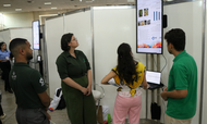 Quatro jovens estão reunidos em um estande de exposição, observando atentamente uma tela vertical que exibe gráficos e informações acadêmicas. Dois rapazes e uma moça vestem uniformes em tons de verde, enquanto outra jovem usa um colete amarelo sobre calça rosa e opera um notebook. O ambiente é amplo, com divisórias brancas e outros painéis digitais ao fundo, sugerindo a realização de um evento científico ou feira de profissões.