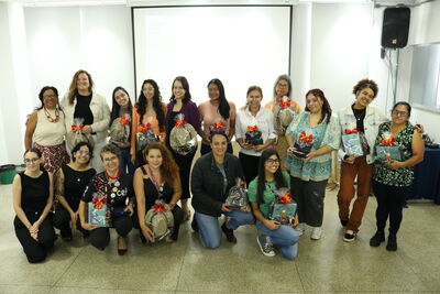 Um grupo de dezenove mulheres posa sorridente em uma sala ampla, segurando kits de presente decorados com laços laranjas. Elas estão distribuídas em duas fileiras, com algumas agachadas à frente e outras em pé ao fundo, vestindo roupas variadas que vão de camisetas casuais a blazers. O ambiente possui paredes brancas, um telão de projeção centralizado e uma caixa de som no canto superior direito.