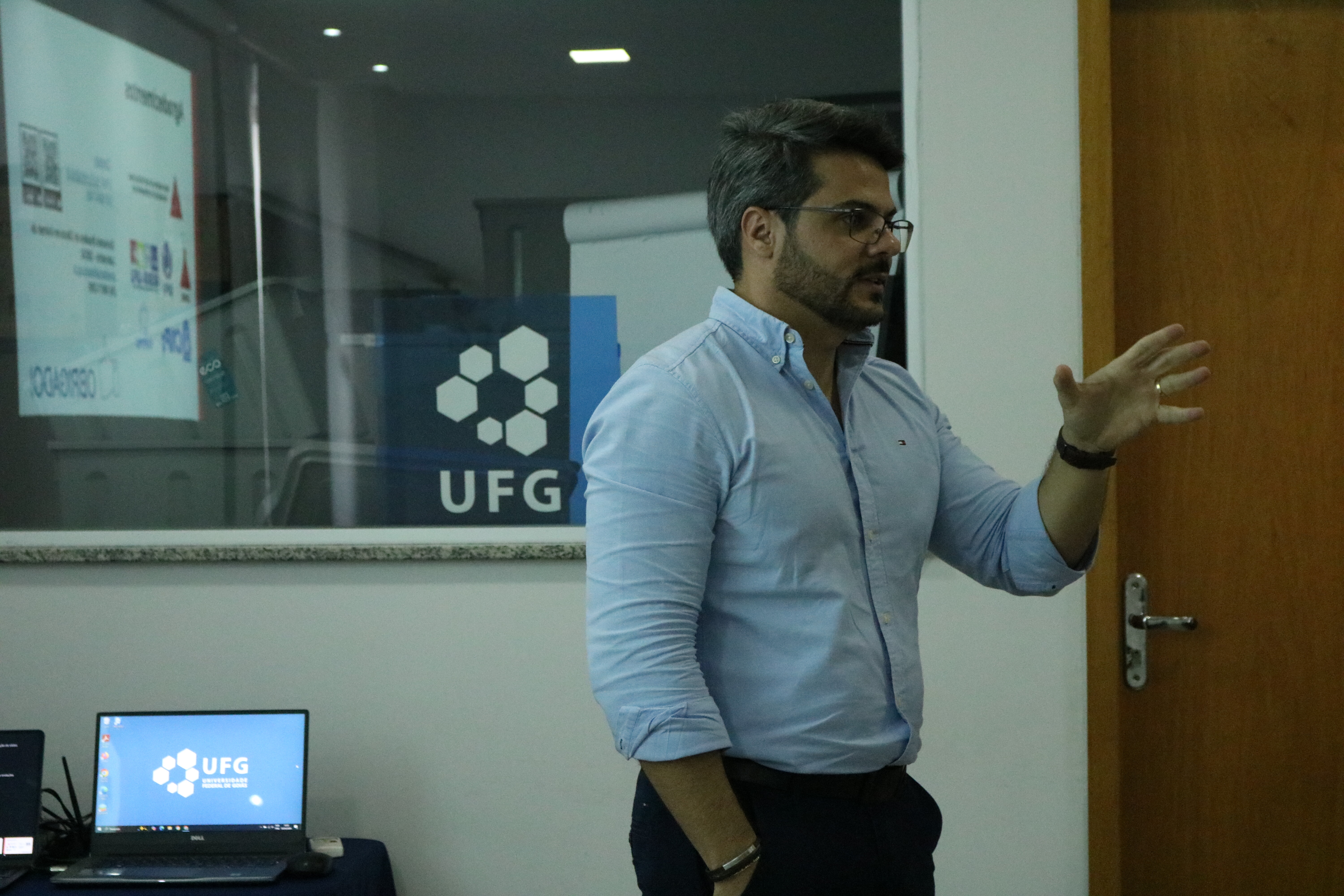 Um homem de barba e óculos, vestindo uma camisa social azul clara, faz uma apresentação em pé, gesticulando com a mão direita. Ao fundo, uma parede branca contém uma janela de vidro que reflete o logotipo da UFG e um cartaz informativo. No canto inferior esquerdo, vê-se um notebook aberto exibindo a marca da universidade na tela, próximo a uma porta de madeira.