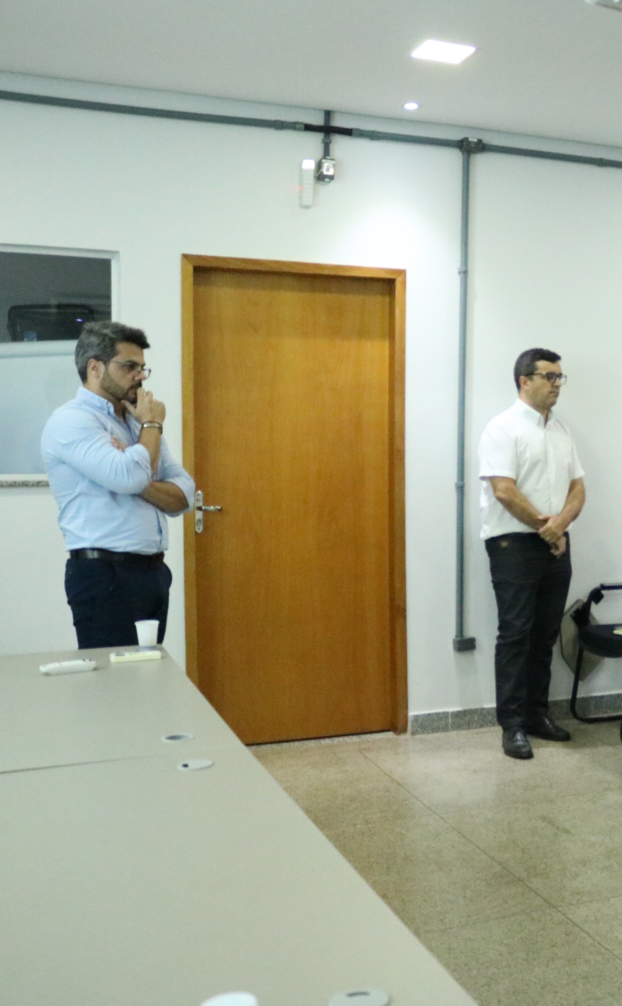 Dois homens estão em pé em uma sala de reuniões clara, posicionados próximos a uma porta de madeira fechada e a uma parede branca. O homem à esquerda veste camisa azul e observa pensativo, enquanto o homem à direita usa camisa branca e mantém as mãos cruzadas à frente do corpo. Em primeiro plano, vê-se parte de uma mesa cinza, sugerindo um ambiente de trabalho ou discussão institucional.