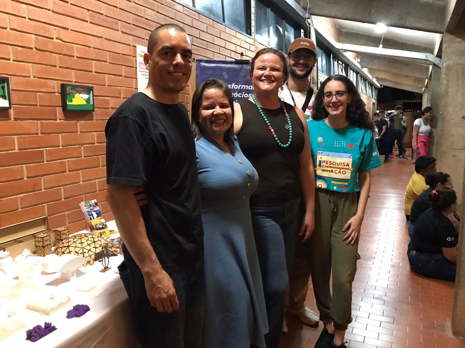 Fotografia colorida em plano médio de um grupo de cinco pessoas (três mulheres e dois homens) posando sorridentes em um corredor interno. Eles estão posicionados ao lado de uma mesa de exposição encostada em uma parede de tijolos aparentes (tijolinho ecológico).