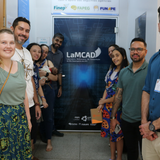 Um grupo de meninas e jovens do movimento escoteiro, vestindo seus uniformes típicos em tons de bege e azul, posa para uma foto em frente ao prédio do LaMCAD na UFG. Elas aparecem sorridentes, acompanhadas por alguns adultos, em uma área externa pavimentada com o céu azul ao fundo. A imagem registra uma visita educativa e de integração entre a comunidade juvenil e o ambiente universitário de tecnologia.