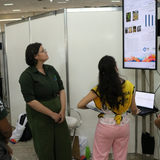 Quatro jovens estão reunidos em um estande de exposição, observando atentamente uma tela vertical que exibe gráficos e informações acadêmicas. Dois rapazes e uma moça vestem uniformes em tons de verde, enquanto outra jovem usa um colete amarelo sobre calça rosa e opera um notebook. O ambiente é amplo, com divisórias brancas e outros painéis digitais ao fundo, sugerindo a realização de um evento científico ou feira de profissões.