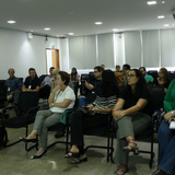 Uma plateia composta por homens e mulheres está sentada em cadeiras pretas dentro de uma sala de reuniões com paredes brancas e iluminação embutida. Os participantes, vestidos com roupas casuais e profissionais, aparecem atentos e voltados para a mesma direção, sugerindo o acompanhamento de uma apresentação. Ao fundo, observam-se janelas com persianas cinzas e uma porta de madeira aberta.