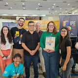 Uma fotografia colorida em plano médio mostra um grupo de seis pessoas (três mulheres e três homens) posando sorridentes em um estande de eventos. O ambiente é um grande pavilhão ou centro de convenções com teto alto e luzes embutidas. Ao fundo, banners e cartazes indicam um evento ligado à UFG