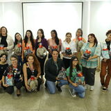 Um grupo de dezenove mulheres posa sorridente em uma sala ampla, segurando kits de presente decorados com laços laranjas. Elas estão distribuídas em duas fileiras, com algumas agachadas à frente e outras em pé ao fundo, vestindo roupas variadas que vão de camisetas casuais a blazers. O ambiente possui paredes brancas, um telão de projeção centralizado e uma caixa de som no canto superior direito.