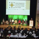 A imagem mostra o interior de um auditório durante o "Evento Integrador do Ciclo 2026 do Programa de Formação em Saúde Brasil-Angola". A fotografia é tirada de trás da plateia, voltada para o palco iluminado onde compõe-se uma mesa de autoridades.