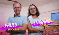 A imagem apresenta Paulo Sucasas e Luciane Costa sorridentes, em pé e de braços cruzados, posicionados lado a lado em um ambiente interno. Seus nomes aparecem em destaque sobre faixas roxas e amarelas à frente de cada um. No canto inferior direito, observa-se o logotipo da Rede Globo, indicando uma participação em programa televisivo.