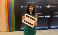 Uma mulher sorridente, de cabelos cacheados e vestido verde, segura um certificado de premiação à frente de um painel preto. O fundo exibe logomarcas de instituições como SEBRAE, SESI, IEL e do Governo Federal, indicando um evento oficial de reconhecimento. À esquerda, nota-se uma coluna decorada com grafismos coloridos que remetem a padrões indígenas ou artísticos.
