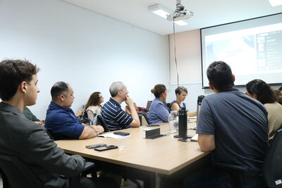Um grupo de nove pessoas está reunido em torno de uma grande mesa de madeira em um escritório com paredes de vidro e persianas. Os participantes, homens e mulheres em trajes casuais e formais, utilizam notebooks e mantêm garrafas de água sobre a mesa durante a conversa. Ao fundo, observa-se uma estante com livros e objetos decorativos, além de uma televisão fixada na parede. A cena registra um momento de trabalho colaborativo e troca de ideias em um ambiente profissional organizado.