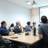 Um grupo de nove pessoas está reunido em torno de uma grande mesa de madeira em um escritório com paredes de vidro e persianas. Os participantes, homens e mulheres em trajes casuais e formais, utilizam notebooks e mantêm garrafas de água sobre a mesa durante a conversa. Ao fundo, observa-se uma estante com livros e objetos decorativos, além de uma televisão fixada na parede. A cena registra um momento de trabalho colaborativo e troca de ideias em um ambiente profissional organizado.