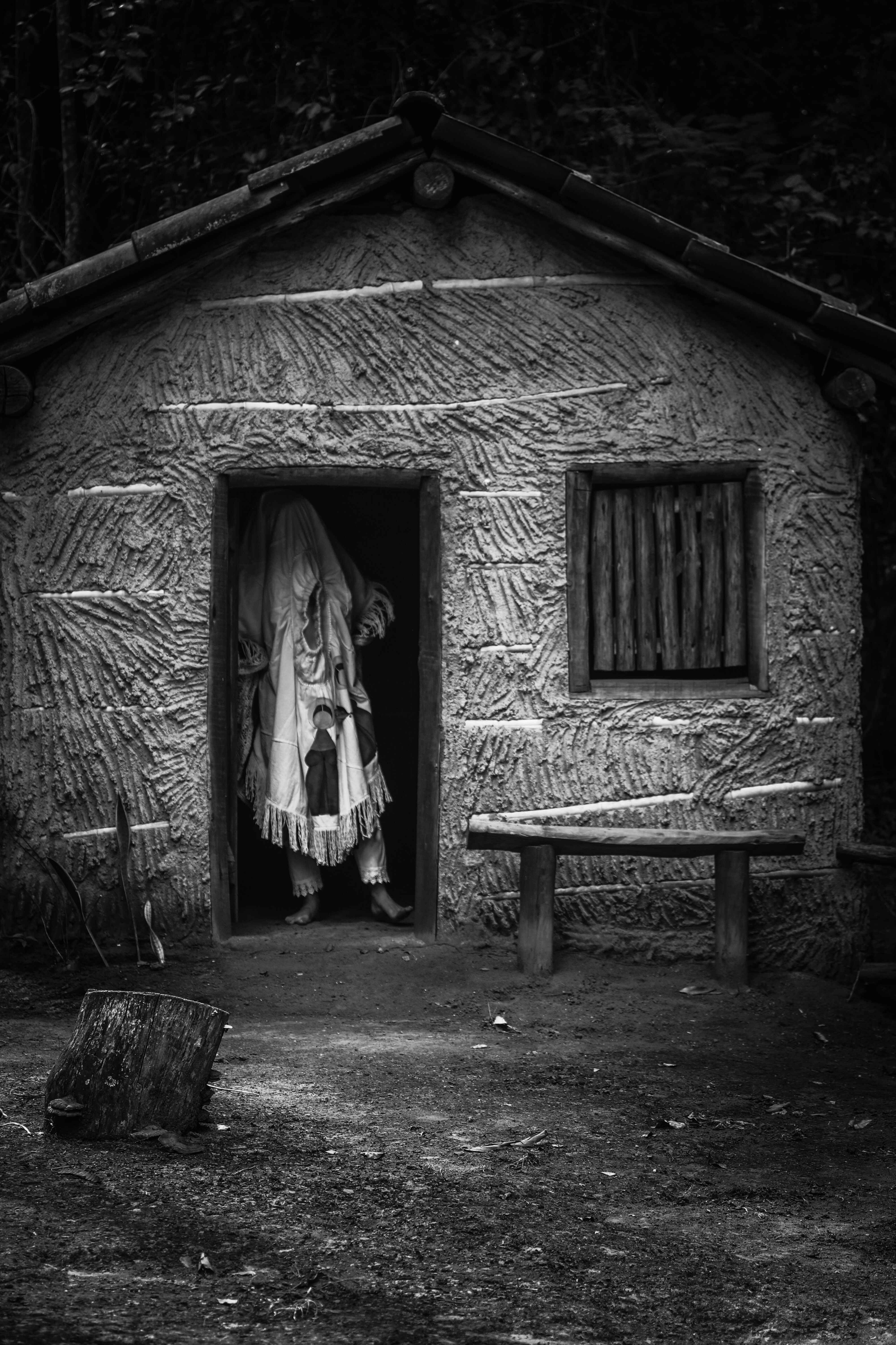 Uma figura humana com o rosto coberto aparece na porta de uma casa de pau à pique.