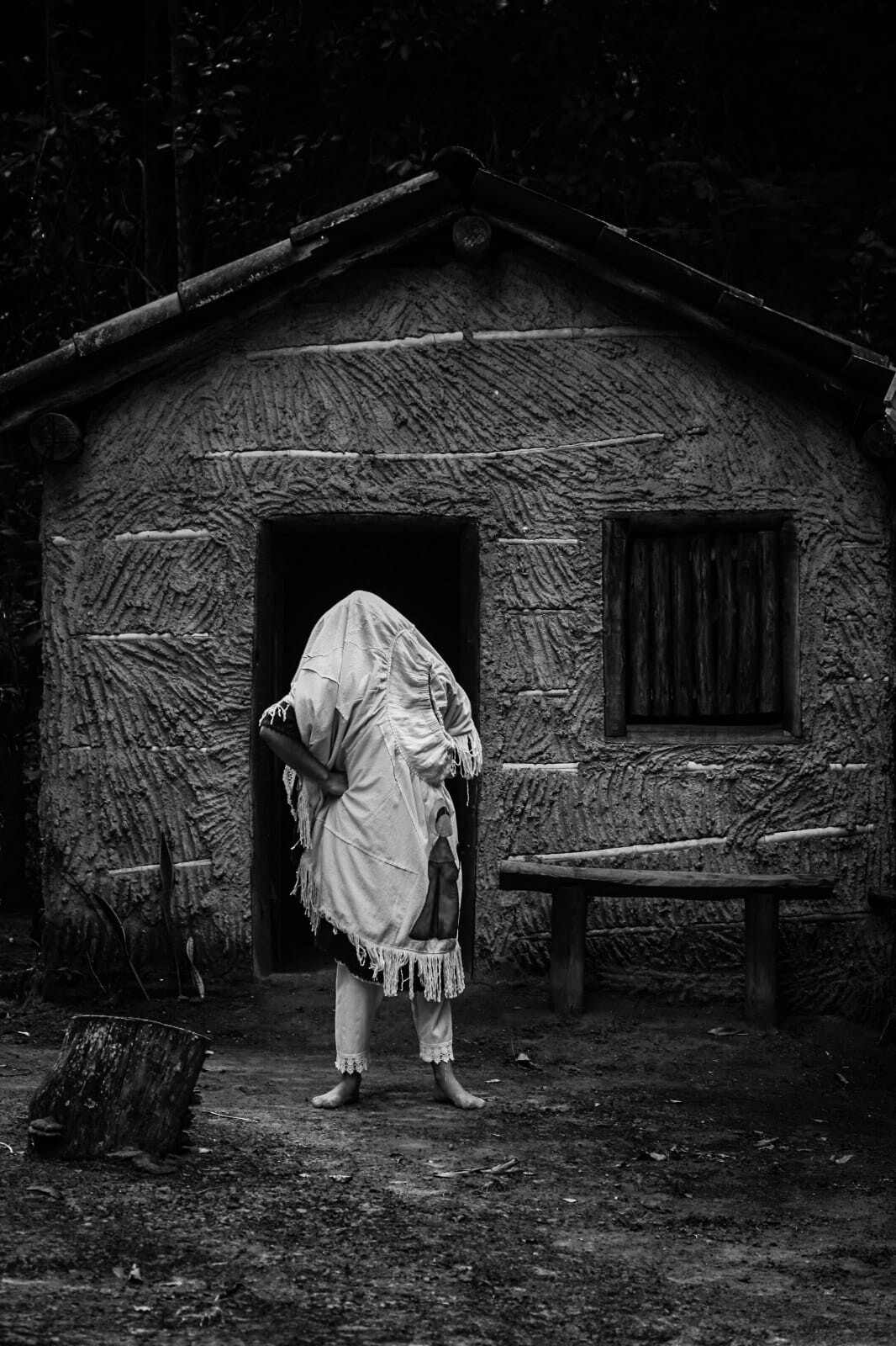 A figura humana que tem o corpo coberto passa pela porta da casa de pau à pique