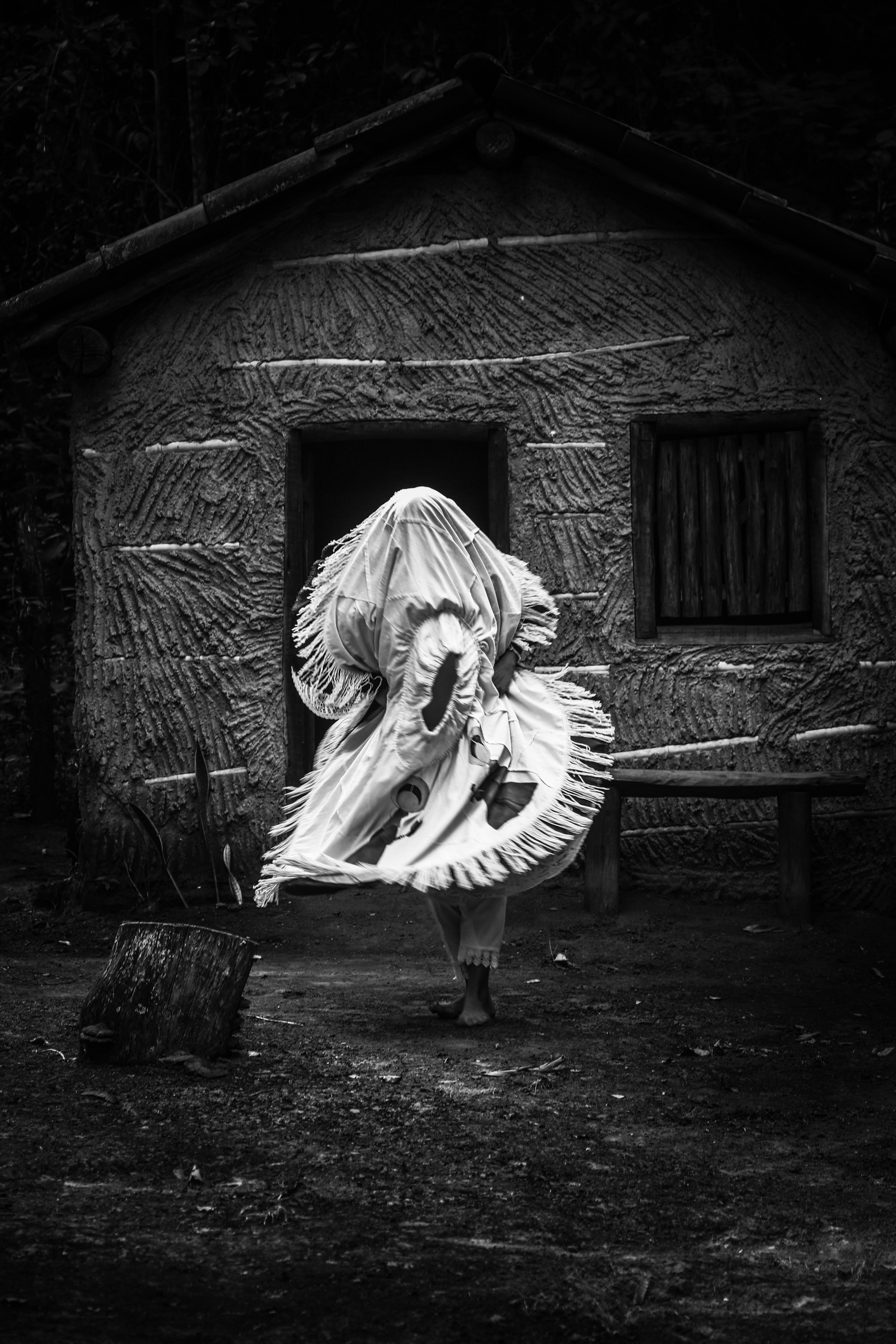 a figura humana que tem o corpo coberto, sai para fora da casa de pau à pique e está em movimento dançante